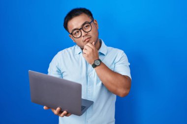 Chinese young man using computer laptop with hand on chin thinking about question, pensive expression. smiling with thoughtful face. doubt concept. 
