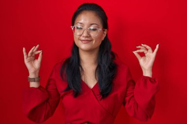 Asian young woman standing over red background relaxed and smiling with eyes closed doing meditation gesture with fingers. yoga concept. 