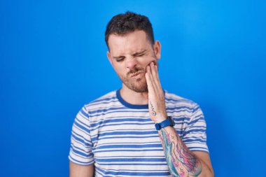 Young hispanic man standing over blue background touching mouth with hand with painful expression because of toothache or dental illness on teeth. dentist 