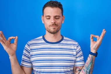 Young hispanic man standing over blue background relaxed and smiling with eyes closed doing meditation gesture with fingers. yoga concept. 