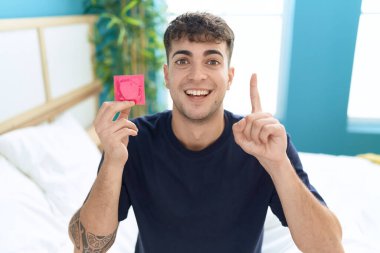 Young hispanic man sitting on the bed holding condom surprised with an idea or question pointing finger with happy face, number one 