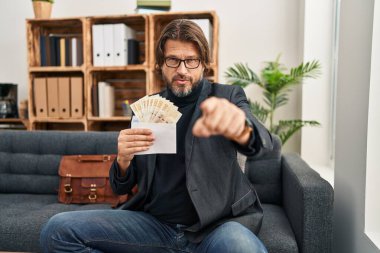 Handsome middle age man at consultation office holding dkk banknotes pointing with finger to the camera and to you, confident gesture looking serious 