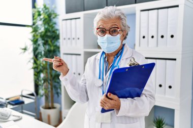 Yaşlı kadın, gri saçlı, doktor üniforması giyen ve güvenlik maskesi takan mutlu bir şekilde elini ve parmağını yana doğru gösteren. 