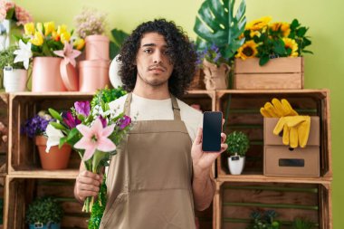 Hispanic man with curly hair working at florist shop showing smartphone screen depressed and worry for distress, crying angry and afraid. sad expression. 