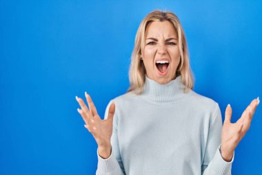 Young caucasian woman standing over blue background crazy and mad shouting and yelling with aggressive expression and arms raised. frustration concept. 