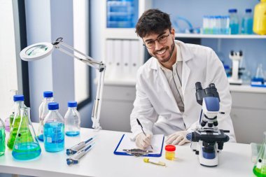 Young hispanic man scientist writing on document holding urine test tube at laboratory