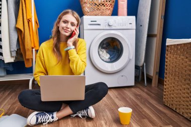 Young blonde woman using laptop talking on smartphone waiting for washing machine at laundry room