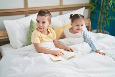 Two kids reading story book sitting on bed at bedroom
