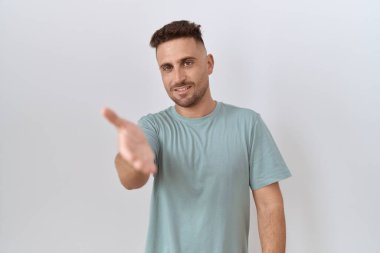 Hispanic man with beard standing over white background smiling cheerful offering palm hand giving assistance and acceptance. 