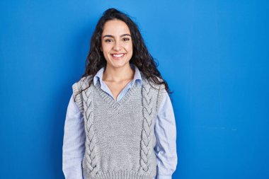 Mavi arka planda, yüzünde mutlu ve havalı bir gülümsemeyle duran esmer bir kadın. şanslı kişi. 