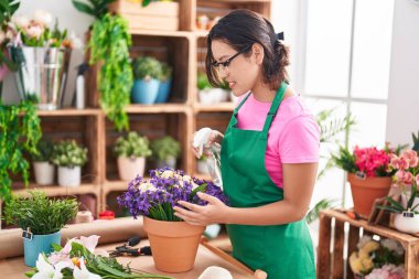 Young hispanic woman florist using diffuser watering plant at florist