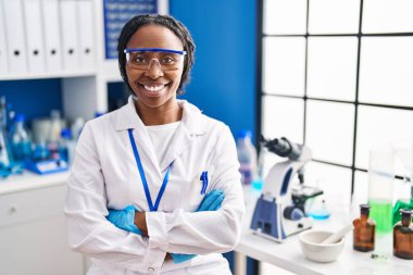 African american woman wearing scientist uniform sitting with arms crossed gesture laboratory