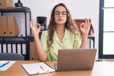 Young hispanic woman working at the office wearing glasses relax and smiling with eyes closed doing meditation gesture with fingers. yoga concept. 