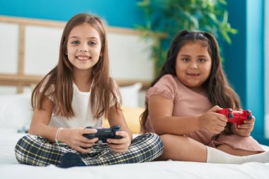 Two kids playing video game sitting on bed at bedroom