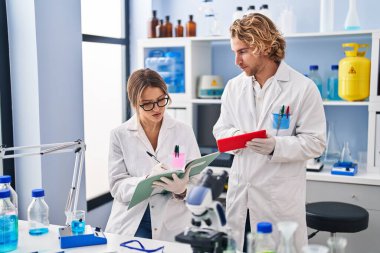 Man and woman wearing scientist uniform writing on notebook using touchpad at laboratory