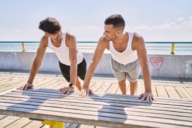 İki İspanyol erkek, deniz kenarında gerinerek gülümsüyor.