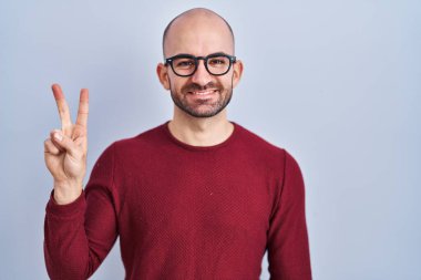 Young bald man with beard standing over white background wearing glasses showing and pointing up with fingers number two while smiling confident and happy. 