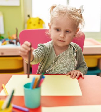 Adorable blonde girl preschool student sitting on table drawing on paper at kindergarten