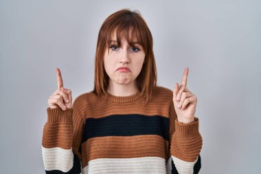 Young beautiful woman wearing striped sweater over isolated background pointing up looking sad and upset, indicating direction with fingers, unhappy and depressed. 