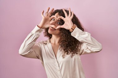 Hispanic woman with curly hair standing over pink background doing heart shape with hand and fingers smiling looking through sign 