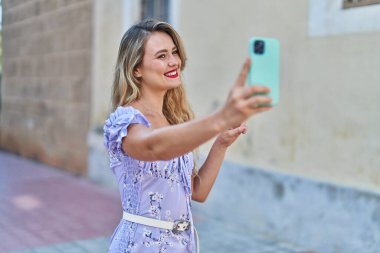 Genç ve güzel İspanyol kadın gülümsüyor. Kendine güveni tam. Sokakta akıllı telefonun yanında selfie çekiyor.