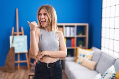 Blonde caucasian woman standing at living room smiling with happy face looking and pointing to the side with thumb up. 