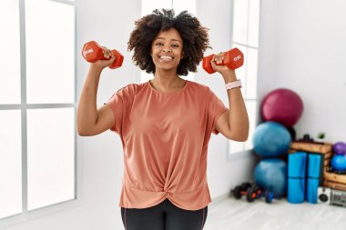 Genç Afrikalı Amerikalı kadın gülümsüyor spor merkezinde halterleri kullanarak kendine güven eğitimi alıyor