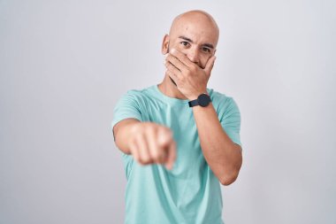 Middle age bald man standing over white background laughing at you, pointing finger to the camera with hand over mouth, shame expression 
