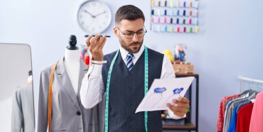 Young hispanic man tailor listening voice message by smartphone reading document at tailor shop