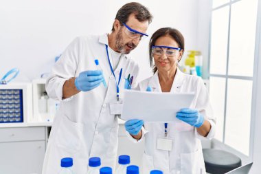 Orta yaşlı erkek ve kadın partnerler bilim adamı üniforması giyip doküman okuyor ve laboratuarda test tüpü tutuyorlar.