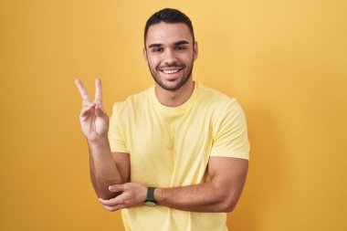 Young hispanic man standing over yellow background smiling with happy face winking at the camera doing victory sign with fingers. number two. 
