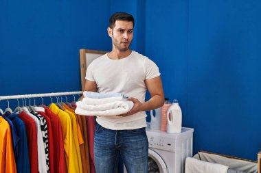 Young hispanic man with beard holding folded clean towels at laundry room in shock face, looking skeptical and sarcastic, surprised with open mouth 