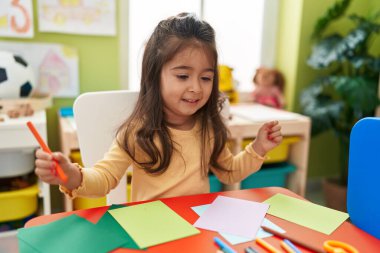Adorable hispanic girl preschool student sitting on table drawing on paper at kindergarten