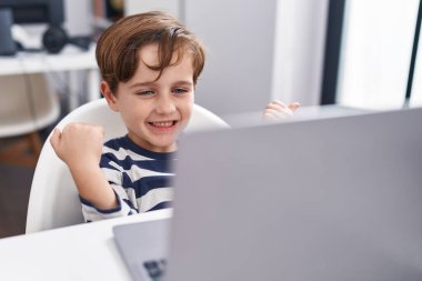 Adorable hispanic boy student using laptop with cheerful expression at classroom