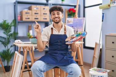 Arab man with beard painter sitting at art studio holding palette smiling happy pointing with hand and finger to the side 