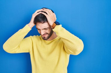Hispanic man standing over blue background suffering from headache desperate and stressed because pain and migraine. hands on head. 