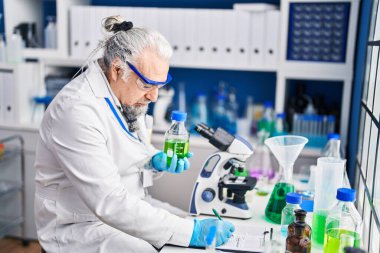 Middle age grey-haired man scientist writing on document holding bottle at laboratory