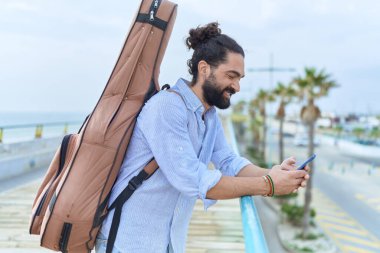Young hispanic man musician using smartphone holding guitar case at seaside
