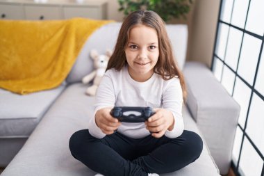 Adorable hispanic girl playing video game sitting on sofa at home