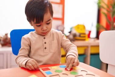 Adorable hispanic boy playing with maths puzzle game sitting on table at kindergarten