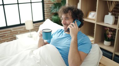 Genç İspanyol adam akıllı telefonda konuşuyor. Yatak odasında kahve içiyor.