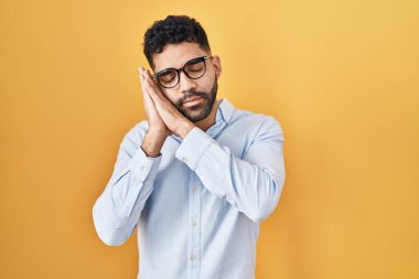 Hispanic man with beard standing over yellow background sleeping tired dreaming and posing with hands together while smiling with closed eyes. 