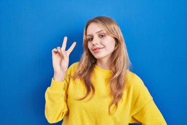 Mavi arka planda dikilmiş, gülümseyen, parmaklarını zafer işareti yaparken gösteren beyaz bir kadın. İki numara.. 