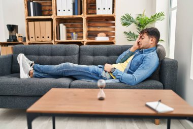 Young hispanic man patient stressed lying on sofa at psychology clinic