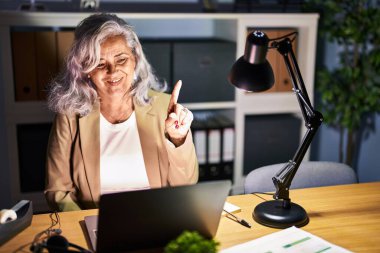 Middle age woman with grey hair working using computer laptop late at night showing and pointing up with finger number one while smiling confident and happy. 