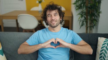 Young hispanic man doing heart gesture sitting on sofa at home