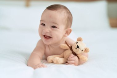 Bebek yatakta uzanmış, yatak odasında oyuncak ayıyı tutuyor.