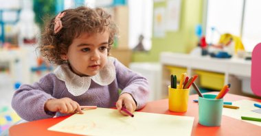Adorable hispanic girl preschool student sitting on table drawing on paper at kindergarten