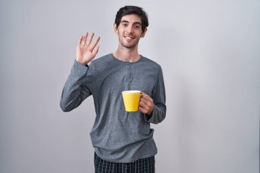 Young hispanic man wearing pajama drinking a cup of coffee waiving saying hello happy and smiling, friendly welcome gesture 