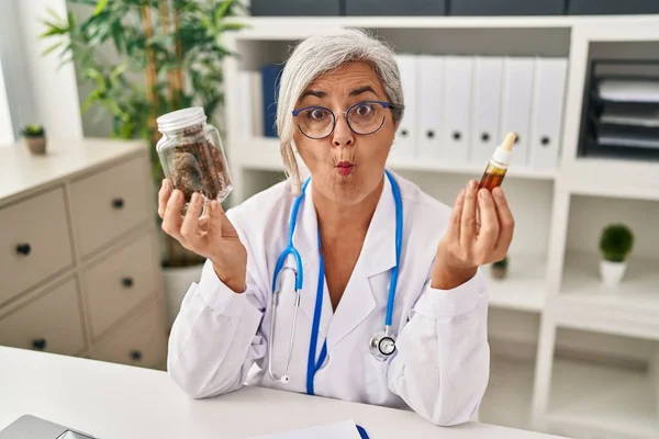 Middle age woman doctor holding cbd oil making fish face with mouth and squinting eyes, crazy and comical.